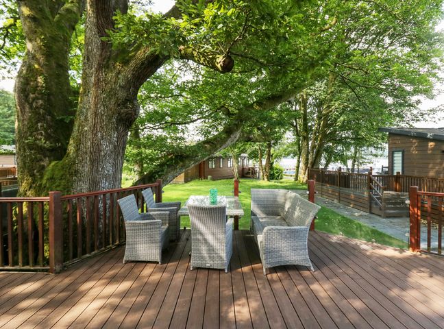 An outdoor wooden deck with wicker chairs and a table surrounded by trees at Tranquil Waters in Lakeside 16 White Cross Bay near Windermere