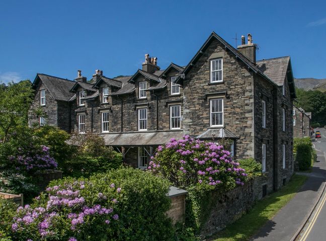 A stone house with multiple chimneys and white framed windows surrounded by green bushes and purple flowers at Waverley in Coniston