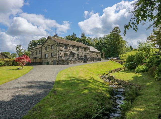 A stone house with a gravel driveway green lawn and a small stream flowing beside it at Fell View House in Lambrigg near Kendal