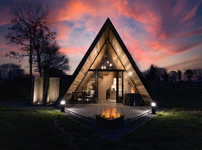 An illuminated triangular cabin with a fire pit in front and trees in the background at The Tri-Pod in Pantcefn near Llanarth