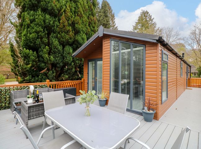 An outdoor deck with a table and chairs and a brown cabin surrounded by trees at Squirrels Nook Waterside Wood 19 White Cross Bay near Windermere