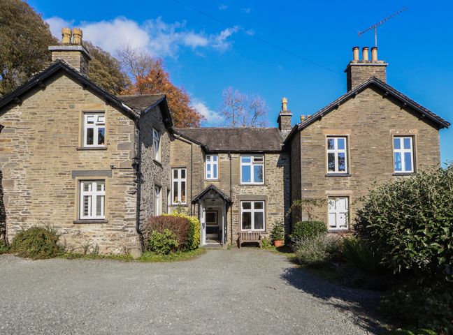 A stone house with multiple windows and chimneys surrounded by plants and gravel driveway at 2 Birkhead Troutbeck near Windermere