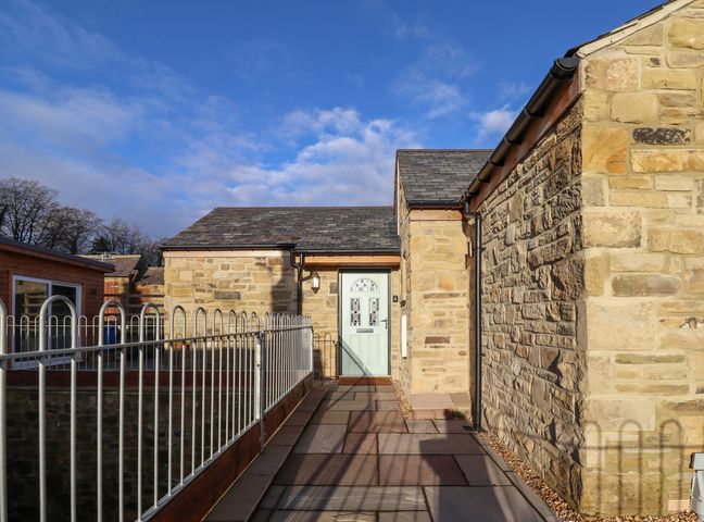 A stone building with a light green door and a metal railing along a walkway at Summerseats Cottage in Alnwick