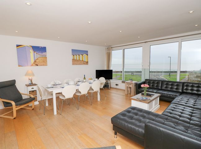 A living room with a black leather sofa a dining table with white chairs a wooden floor and large windows overlooking a road and grassy area at Sea House in Rustington