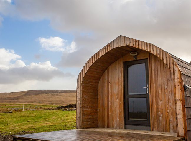 A wooden pod with a glass door on a deck in a grassy landscape at Heisgeir Sollas Isle Of North Uist