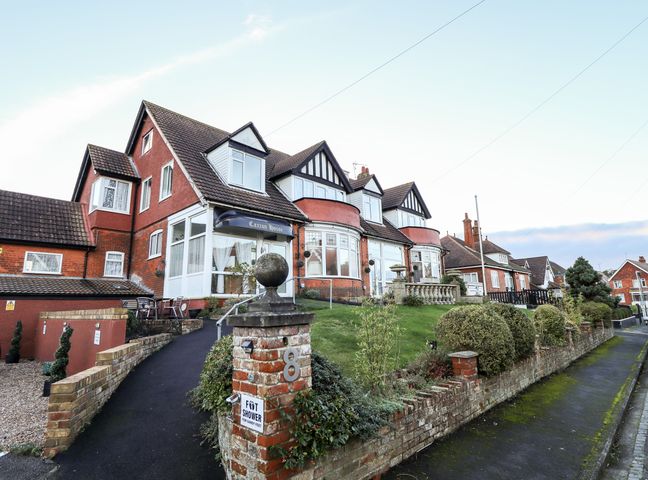 A red brick building with multiple windows and a sloped roof with a sign saying Caxton House and a garden with bushes in Skegness
