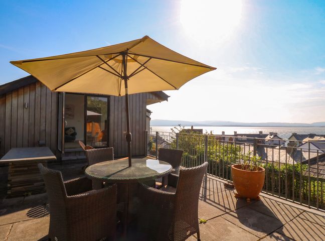 A patio with wicker chairs and a glass table under a large umbrella overlooking a body of water at 1 Gibraltar Terrace in Aberdovey