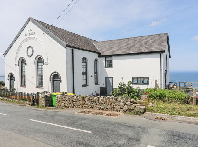 A white building with arched windows and a slate roof by a roadside with a stone wall and ocean in the background at Capel Bethania in Nefyn