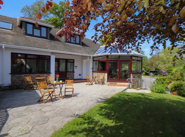 An outdoor patio with wooden tables and chairs in front of a house with a conservatory at Lakeside in Falmouth