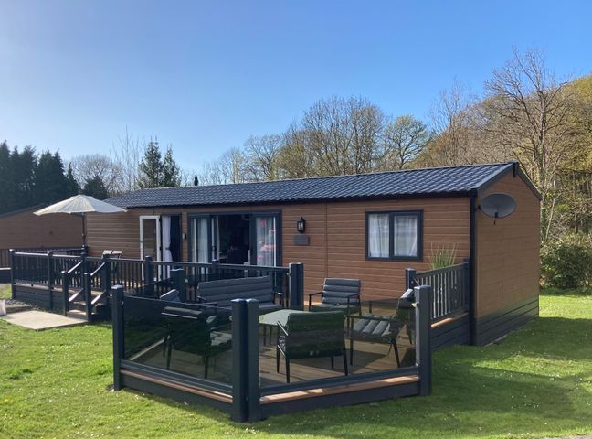 A wooden cabin with black railing deck chairs and a table on a grassy area at 43 The Deri in Llanarth