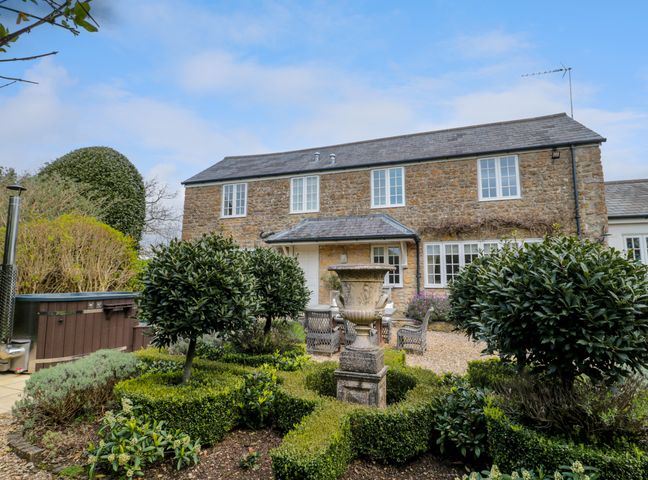 A stone house with white framed windows garden with trimmed bushes and an outdoor seating area at Pomme Blushe House in Shipton Gorge