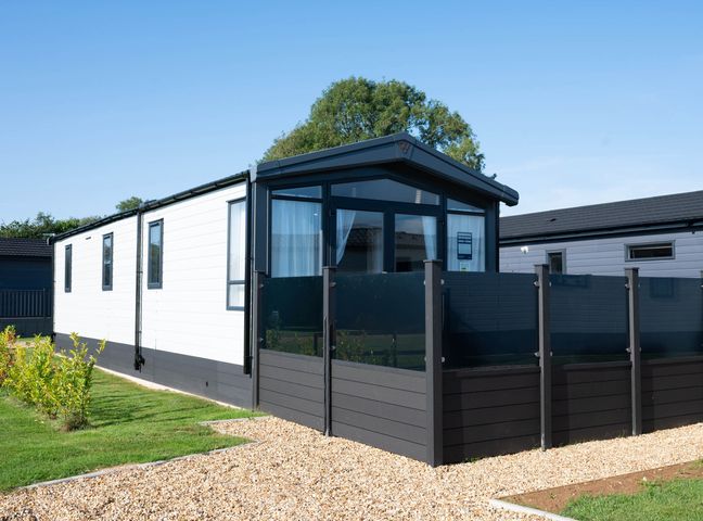 A modern static caravan with black trim and a glass panel fence on a gravel pathway surrounded by grass at Tiggywinkles in Chipping Norton