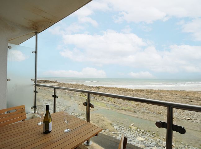 A balcony with a wooden table a bottle and two glasses overlooking a rocky beach and the sea at 2 Horizon View in Westward Ho
