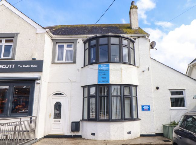 The exterior of a two-story white building with black-framed bay windows and signs for Niles Apartment and parking at Flat 1 Niles Place in St Merryn