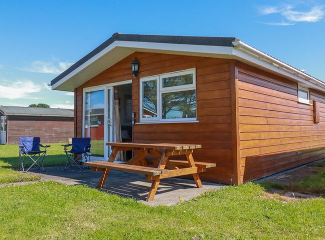 A wooden cabin with a picnic table and two camping chairs outside on a grassy area at Cornish Holiday at Atlantic Bays in St Merryn