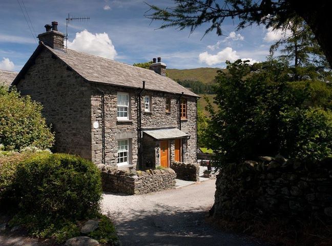 A stone cottage with two wooden doors surrounded by greenery and a stone wall at The Cosy Peacock in Troutbeck
