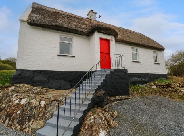 A white cottage with a thatched roof and a red door accessed by stairs surrounded by rocky ground at Aggrafard in Oughterard County Galway