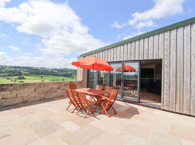 An outdoor patio with wooden table and six chairs under red umbrella adjacent to sliding glass doors and countryside view at Pit Close in Dalwood