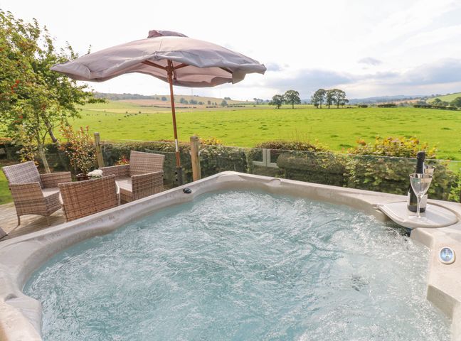 An outdoor hot tub with bubbling water next to a patio table with four chairs and a large umbrella overlooking a green field at Plot 32 High Hesket near Armathwaite