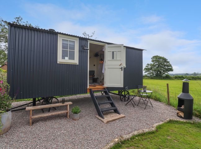 A small black corrugated metal shepherd hut with open door on a gravel area with bench table and chairs and fire pit at La'al Ling in Penrith