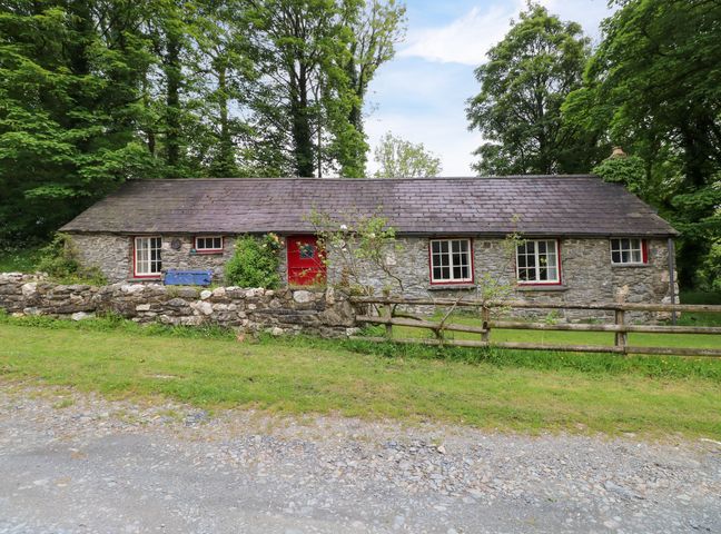 A stone cottage with a red door and windows surrounded by trees and a wooden fence at Penyrallt Fach Cottage in Llandysul