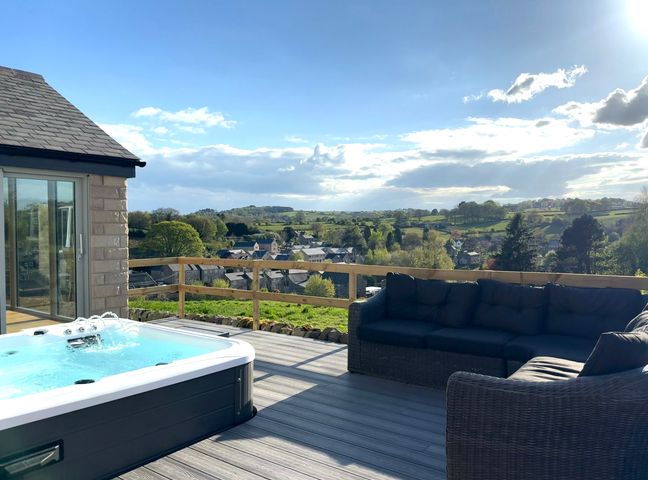 An outdoor deck with a hot tub and wicker sofa overlooking a village and green hills at Barncroft in Tansley near Matlock