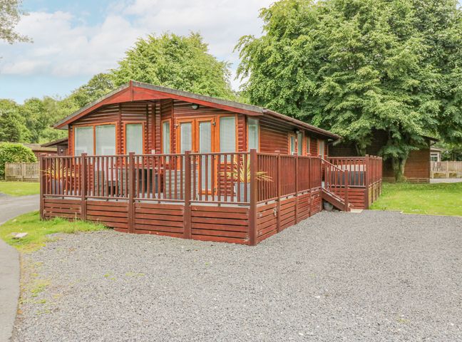 A wooden lodge with a fenced deck surrounded by gravel and trees at Don's Lodge in Grasmere 3 White Cross Bay near Windermere