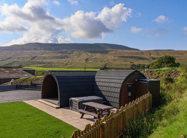 An outdoor area with a curved cabin a picnic table and a fence with hills in the background at Lapwing in Chapel Le Dale near Ingleton