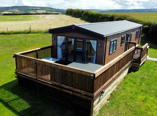 A wooden cabin with a fenced deck surrounded by grass and fields at Aspen Lodge in Whitby