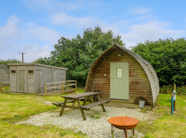 A wooden pod with a picnic table and fire pit outside near wooden sheds at Badger Set @ Penbugle Organic Farm in St Keyne near Duloe