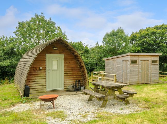 A wooden outdoor pod with a green door beside a wooden shed and a picnic table on gravel at Rabbit Warren Penbugle Organic Farm in St Keyne near Duloe
