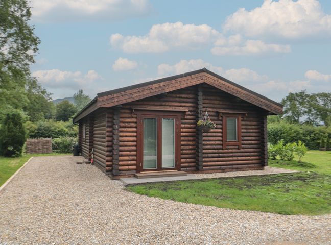 A wooden log cabin with small windows and a gravel driveway surrounded by grass and trees at Coldmoor Lodge 6 in Stokesley