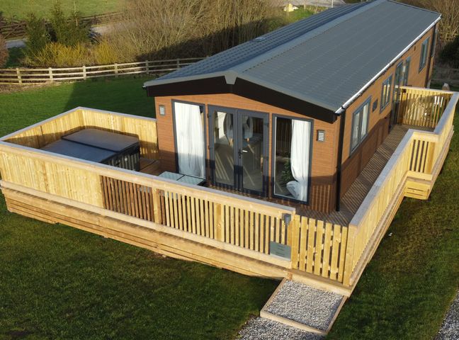 A single-story wooden lodge with a fenced wooden deck and a hot tub on the deck at Juniper Lodge in Whitby