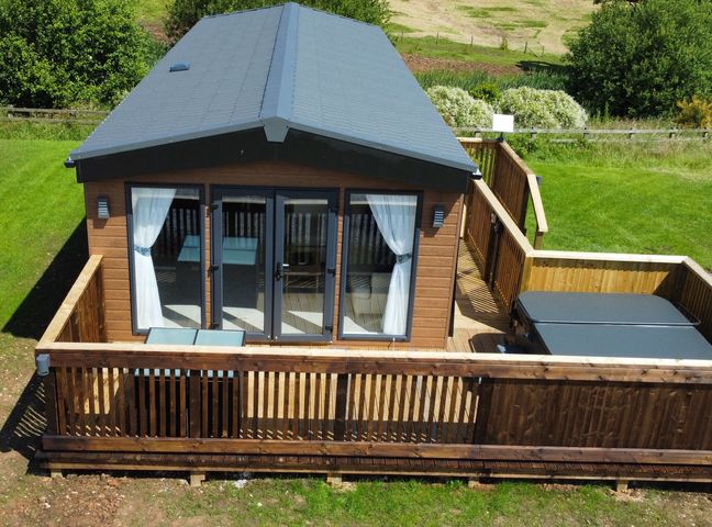A small wooden cabin with glass doors on a wooden deck with patio furniture and a covered hot tub in a grassy area at Willow Lodge in Whitby