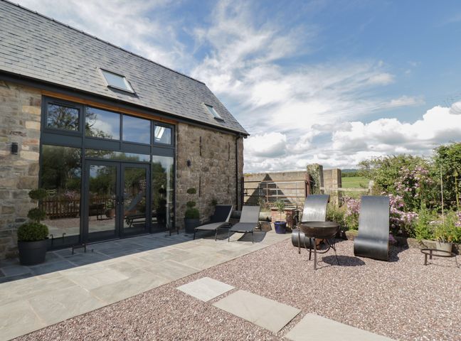 A stone building with large glass doors and windows beside a patio with lounge chairs and metal fire pit at The Hay Barn in Huntley