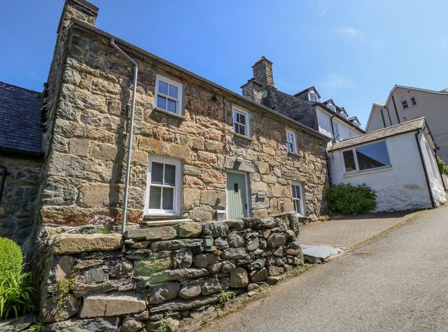 A stone exterior cottage with windows a door and a stone wall next to a paved driveway at Caereinion in Harlech