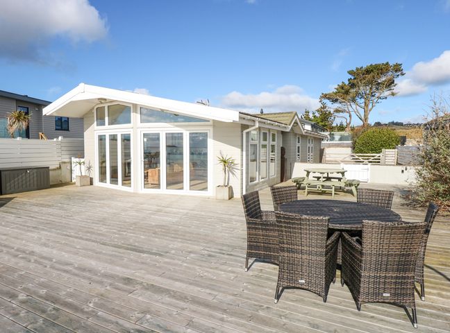 An outdoor wooden deck with wicker chairs around a table and a wooden picnic table near a building at The Warren M45 in Abersoch