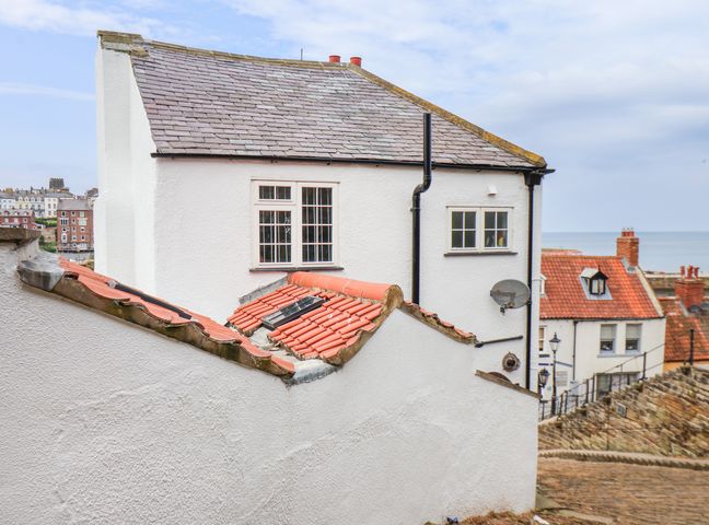 A white building with tiled roofs and windows near a cobblestone street overlooking other buildings and the sea at Steps Cottage in Whitby