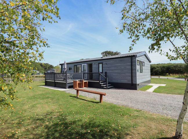 An exterior view of a single-story cabin with a deck and outdoor seating surrounded by grass and trees at Sunset Lodge in Woodhall Spa