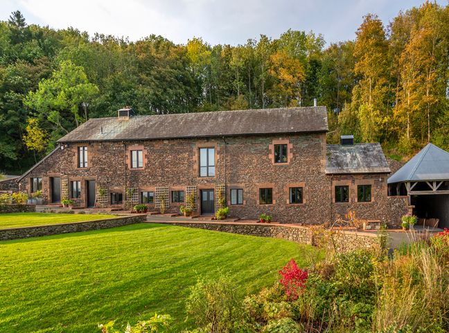 A large stone house with multiple windows and a slate roof in front of a lawn and trees at Bank Barn Wreay Watermillock