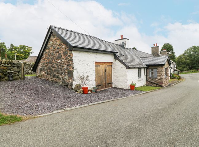 A stone and white painted cottage with wooden double doors and potted plants beside a road at Penrallt in Rhydlydan