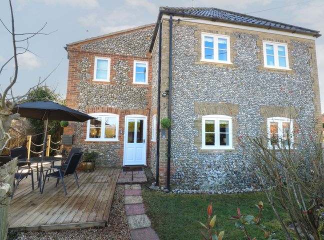Exterior view of a stone cottage with white framed windows a wooden deck with patio table chairs and umbrella at Cobble Cottage in Great Ryburgh
