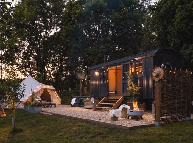An outdoor camping setup with a tent, a small cabin, lounge chairs, a fire pit, and trees at Moonshine in St Erme near Truro