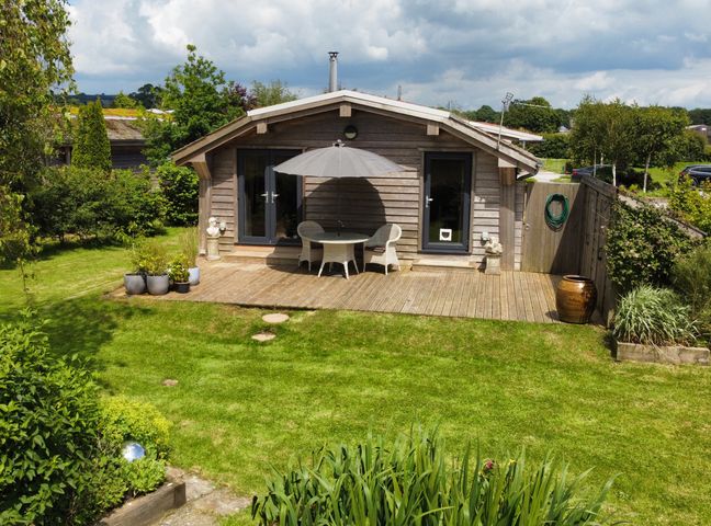 A wooden cabin with a deck patio table umbrella and chairs in a garden at Artisan Lodge in Longridge
