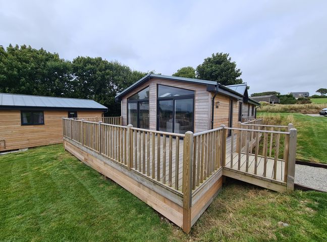 A wooden house with large windows and a fenced wooden deck surrounded by grass at Cove Lodge in Ilfracombe