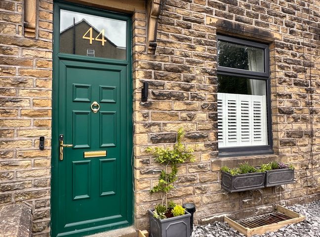 A green front door with number 44 next to a window with white shutters and flower boxes on a stone house wall at Wildfell Cottage in Oxenhope