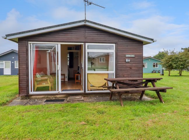 A small wooden cabin with sliding glass doors and a picnic table outside on grass at Pepper Cove near St Merryn