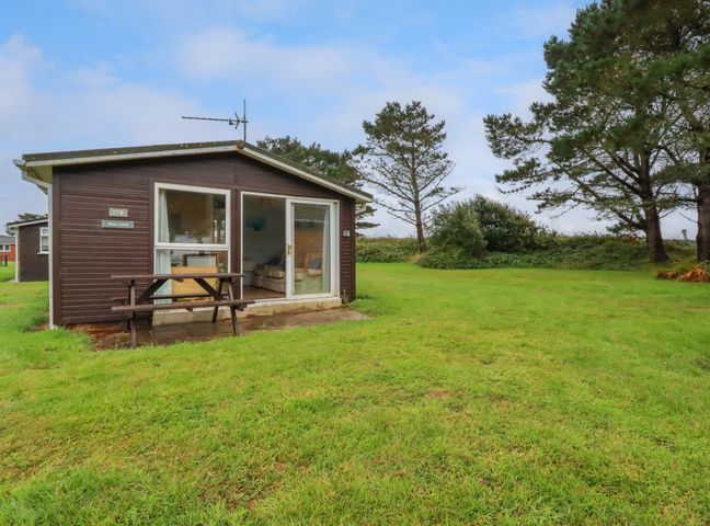 A small brown cabin with a picnic table outside on grass near trees at High Cove near St Merryn