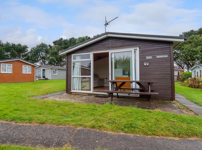 A small dark brown wooden cabin with sliding glass doors and a picnic bench outside at Hawker's Cove near St Merryn
