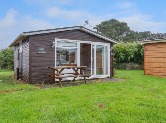 A small wooden cabin with sliding glass doors and a picnic table on a grassy lawn at Newtrain Bay near St Merryn